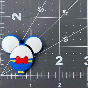Puede incluir: Encanto de goma azul y blanco con forma de cabeza de Mickey Mouse. El encanto tiene una pajarita roja y un bot&oacute;n amarillo.