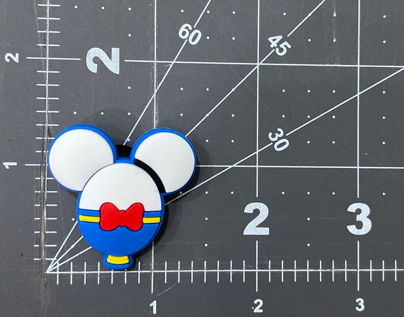 Puede incluir: Encanto de goma azul y blanco con forma de cabeza de Mickey Mouse. El encanto tiene una pajarita roja y un bot&oacute;n amarillo.