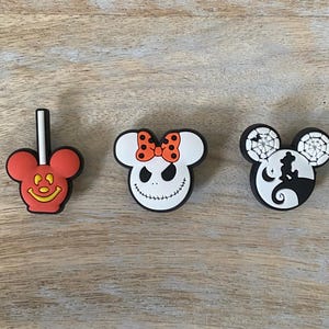 May include: Three colorful shoe charms featuring iconic cartoon characters. One is a red Mickey Mouse lollipop, another is a white Jack Skellington face with an orange bow, and the third is a black and white design with a silhouette.
