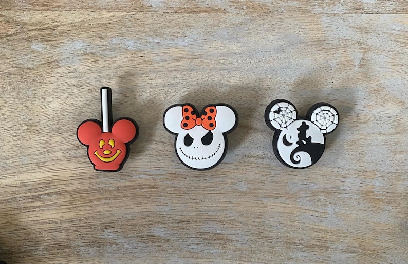 May include: Three colorful shoe charms featuring iconic cartoon characters. One is a red Mickey Mouse lollipop, another is a white Jack Skellington face with an orange bow, and the third is a black and white design with a silhouette.
