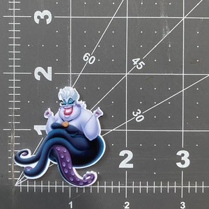 K&ouml;nnte beinhalten: Ein lila und schwarzer Ursula-Aufkleber aus dem Disney-Film Die kleine Meerjungfrau. Ursula sitzt auf ihren Krakenarmen mit erhobenen Armen in einer dramatischen Pose. Sie tr&auml;gt ein lila Kleid und eine Krone auf dem Kopf.