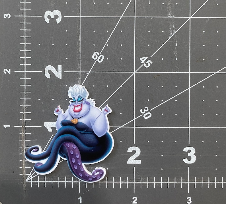 K&ouml;nnte beinhalten: Ein lila und schwarzer Ursula-Aufkleber aus dem Disney-Film Die kleine Meerjungfrau. Ursula sitzt auf ihren Krakenarmen mit erhobenen Armen in einer dramatischen Pose. Sie tr&auml;gt ein lila Kleid und eine Krone auf dem Kopf.