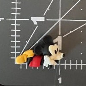 Puede incluir: Una peque&ntilde;a figura tridimensional de Mickey Mouse. La figura est&aacute; tumbada sobre una superficie gris. Mickey Mouse lleva sus caracter&iacute;sticos pantalones cortos rojos, zapatos amarillos y guantes blancos. Su cabeza y orejas son negras y su cara es blanca.