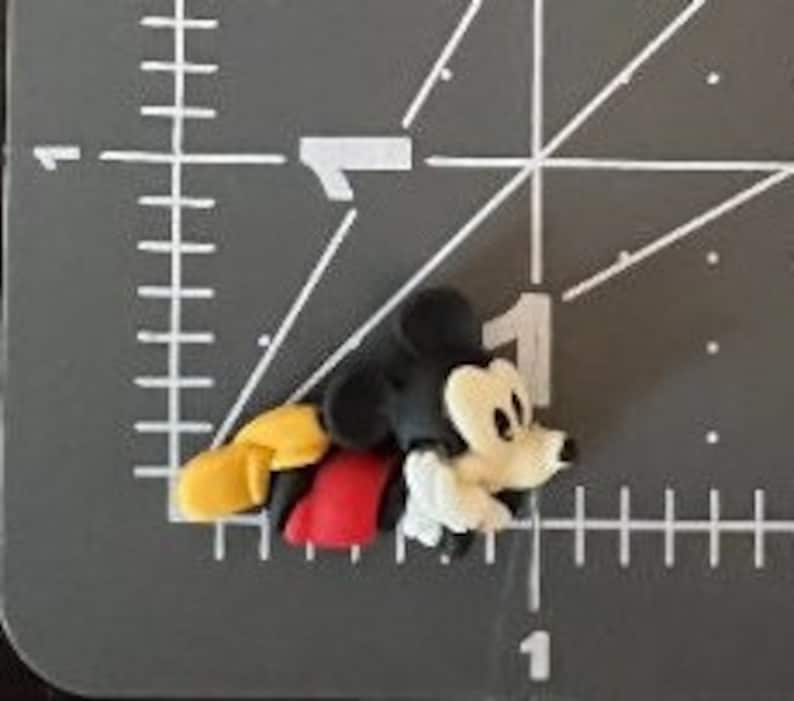 Puede incluir: Una peque&ntilde;a figura tridimensional de Mickey Mouse. La figura est&aacute; tumbada sobre una superficie gris. Mickey Mouse lleva sus caracter&iacute;sticos pantalones cortos rojos, zapatos amarillos y guantes blancos. Su cabeza y orejas son negras y su cara es blanca.