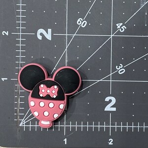 Puede incluir: Encanto de personaje de Minnie Mouse rosa y negro con lunares blancos y un lazo rosa.