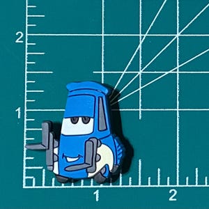 May include: A blue cartoon car with a smiling face and large eyes. The car has a white tire and a black bumper. The car is a character from the animated movie Cars.