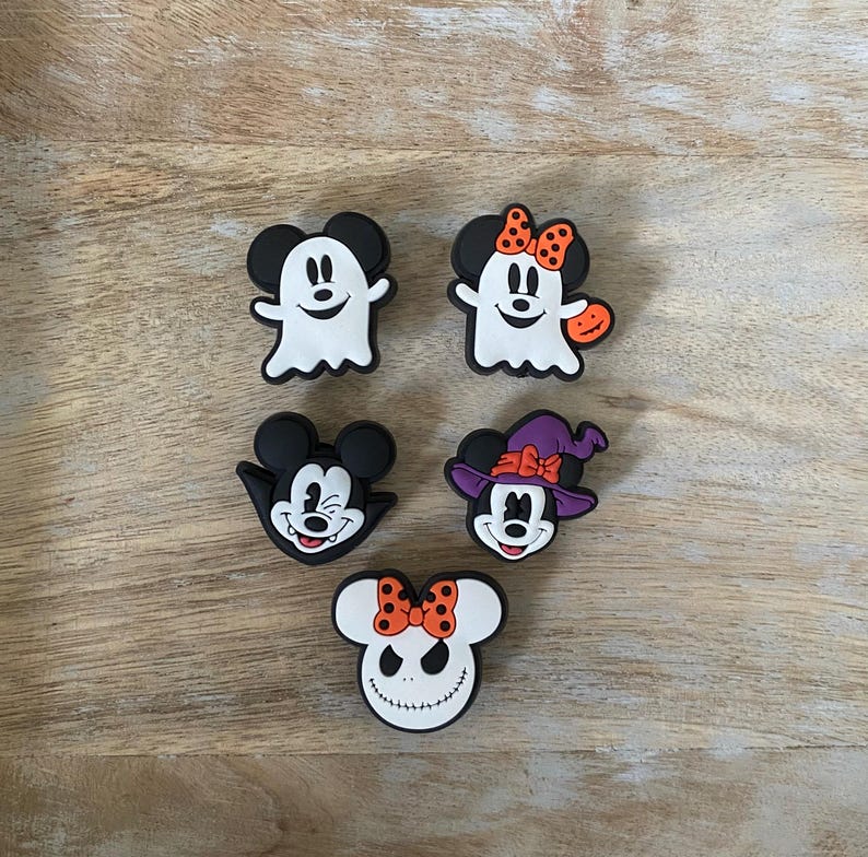 May include: Five Halloween-themed shoe charms featuring cartoon characters. The charms include a ghost, a witch, and a character with a pumpkin. The charms are black, white, orange, and purple, and are arranged on a wooden surface.