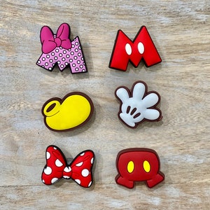 Op de afbeelding: Een verzameling kleurrijke schoenbedels met iconische Disney-ontwerpen. Inclusief een roze polka-dot strik, een rode "M" met witte cirkels, een gele schoen, een witte handschoen, een rode polka-dot strik en een rode Mickey Mouse-kop.