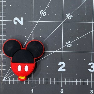 Puede incluir: Un amuleto de goma con forma de Mickey Mouse de color rojo, negro y blanco. El amuleto mide aproximadamente 5 cm de alto y tiene un peque&ntilde;o agujero en la parte superior para sujetarlo a un llavero u otro objeto.