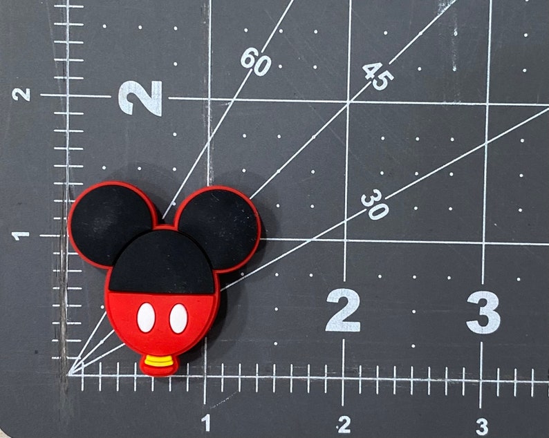 Puede incluir: Un amuleto de goma con forma de Mickey Mouse de color rojo, negro y blanco. El amuleto mide aproximadamente 5 cm de alto y tiene un peque&ntilde;o agujero en la parte superior para sujetarlo a un llavero u otro objeto.