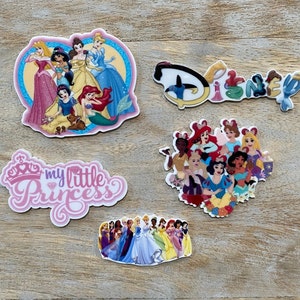 May include: A collection of colorful Disney princess-themed stickers. The stickers feature various princesses, the Disney logo, and the phrase "my little Princess". The stickers are on a wooden surface.