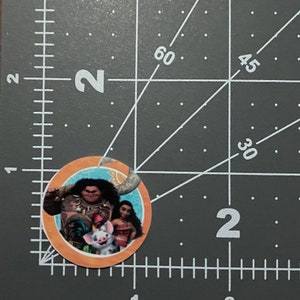 May include: A round sticker featuring characters from the animated movie Moana. The sticker shows Maui, Moana, and Pua in a tropical setting. The sticker is orange and blue.