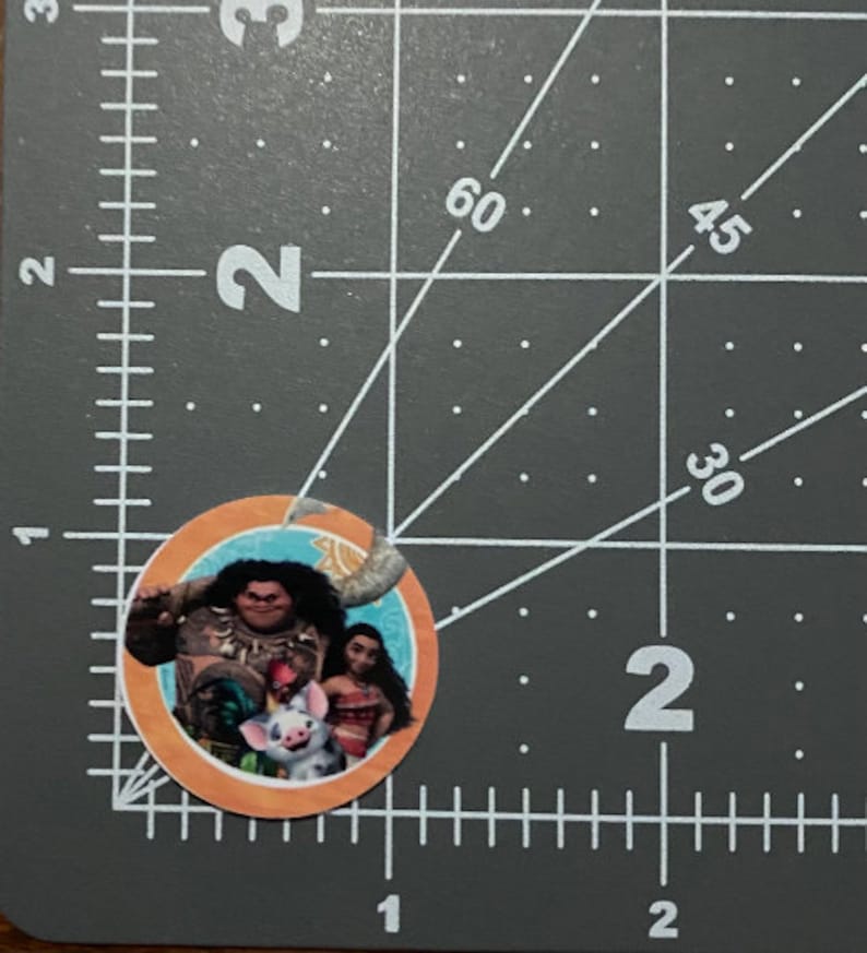 May include: A round sticker featuring characters from the animated movie Moana. The sticker shows Maui, Moana, and Pua in a tropical setting. The sticker is orange and blue.