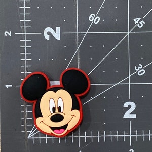 Puede incluir: Un amuleto de cabeza de Mickey Mouse de dibujos animados de color rojo, negro y marr&oacute;n. El amuleto est&aacute; hecho de goma y tiene un acabado brillante.