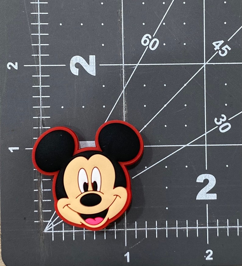 Puede incluir: Un amuleto de cabeza de Mickey Mouse de dibujos animados de color rojo, negro y marr&oacute;n. El amuleto est&aacute; hecho de goma y tiene un acabado brillante.