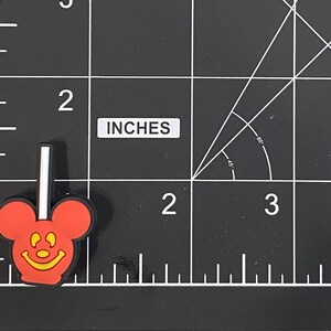 K&ouml;nnte beinhalten: Ein roter und schwarzer Mickey Mouse-f&ouml;rmiger Lutscher-Charm mit einem wei&szlig;en Stiel. Der Charm hat ein gelbes l&auml;chelndes Gesicht. Der Charm ist etwa 2,5 cm hoch.