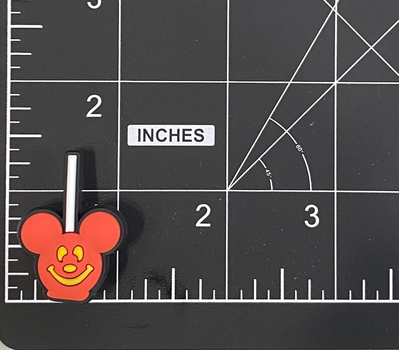 K&ouml;nnte beinhalten: Ein roter und schwarzer Mickey Mouse-f&ouml;rmiger Lutscher-Charm mit einem wei&szlig;en Stiel. Der Charm hat ein gelbes l&auml;chelndes Gesicht. Der Charm ist etwa 2,5 cm hoch.