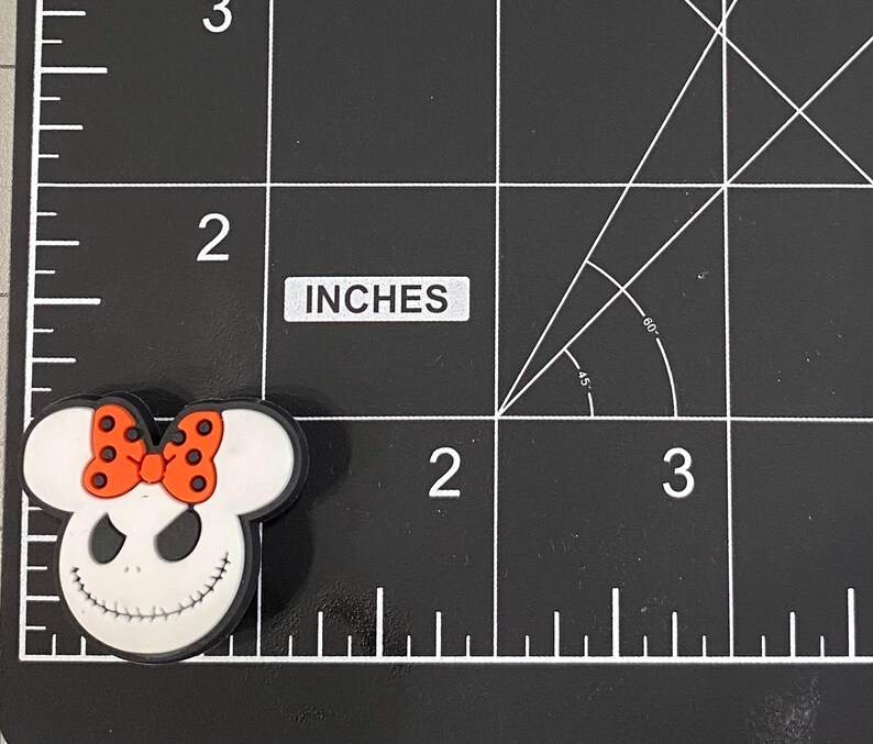 May include: A white and black shoe charm featuring a smiling character face with a black mouth and eyes, and a red bow with black polka dots. The charm is approximately 2 inches in length.