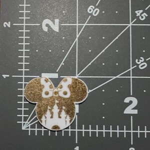 Puede incluir: Una cabeza de rat&oacute;n blanca y dorada con purpurina con un dise&ntilde;o de castillo dentro de las orejas. La cabeza del rat&oacute;n est&aacute; sobre una superficie gris con una regla.