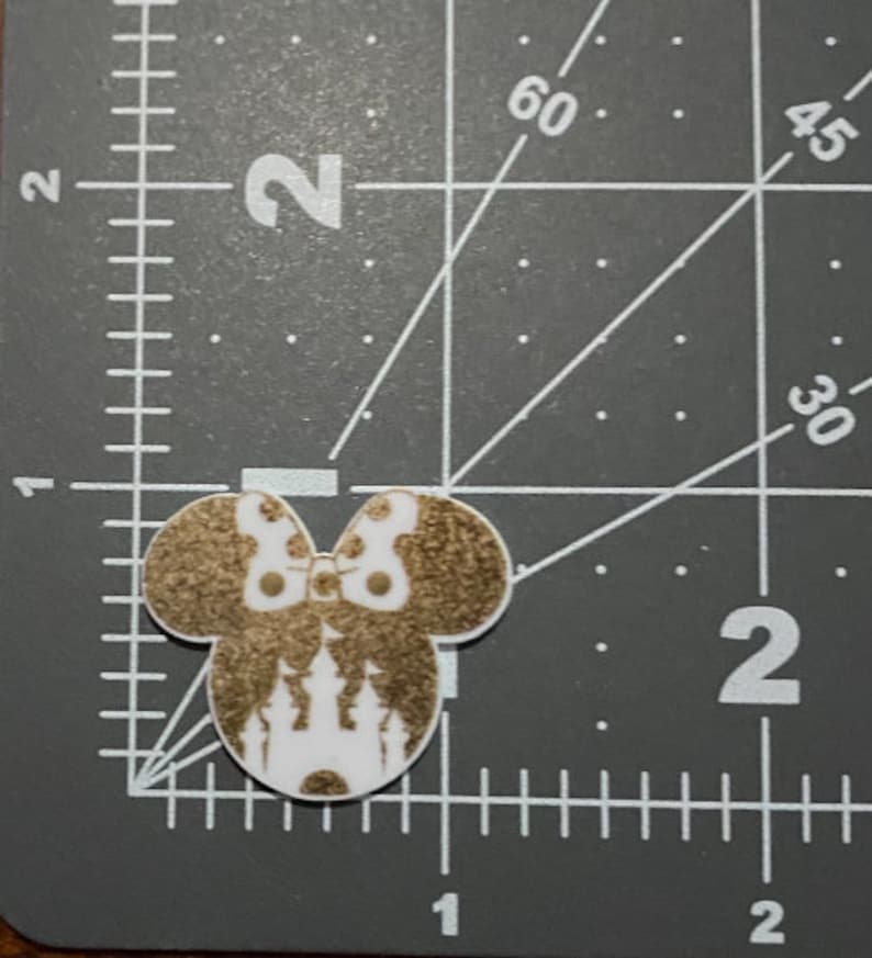 Puede incluir: Una cabeza de rat&oacute;n blanca y dorada con purpurina con un dise&ntilde;o de castillo dentro de las orejas. La cabeza del rat&oacute;n est&aacute; sobre una superficie gris con una regla.