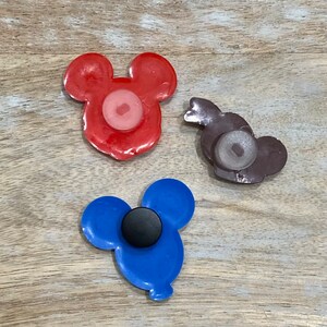 Puede incluir: Tres imanes con forma de cabeza de Mickey Mouse de diferentes colores. El im&aacute;n rojo tiene un centro rosa, el im&aacute;n azul tiene un centro negro y el im&aacute;n marr&oacute;n tiene un centro blanco.