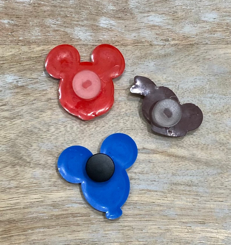 Puede incluir: Tres imanes con forma de cabeza de Mickey Mouse de diferentes colores. El im&aacute;n rojo tiene un centro rosa, el im&aacute;n azul tiene un centro negro y el im&aacute;n marr&oacute;n tiene un centro blanco.