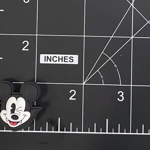 K&ouml;nnte beinhalten: Schwarz-wei&szlig;er Mickey Mouse-f&ouml;rmiger Charm mit zwinkerndem Gesichtsausdruck und roter Zunge. Der Charm ist etwa 3,8 cm lang. Der Charm befindet sich auf einer schwarzen Oberfl&auml;che mit wei&szlig;en Gitterlinien.