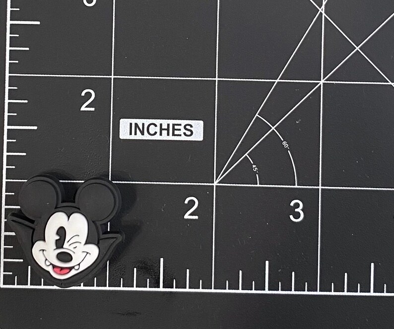 K&ouml;nnte beinhalten: Schwarz-wei&szlig;er Mickey Mouse-f&ouml;rmiger Charm mit zwinkerndem Gesichtsausdruck und roter Zunge. Der Charm ist etwa 3,8 cm lang. Der Charm befindet sich auf einer schwarzen Oberfl&auml;che mit wei&szlig;en Gitterlinien.