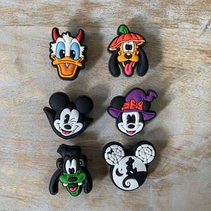 May include: Six colorful cartoon character shoe charms. The charms feature Donald Duck with devil horns, Pluto in a pumpkin hat, Mickey Mouse as a vampire, Minnie Mouse in a witch hat, Goofy, and Mickey Mouse with a Halloween design.
