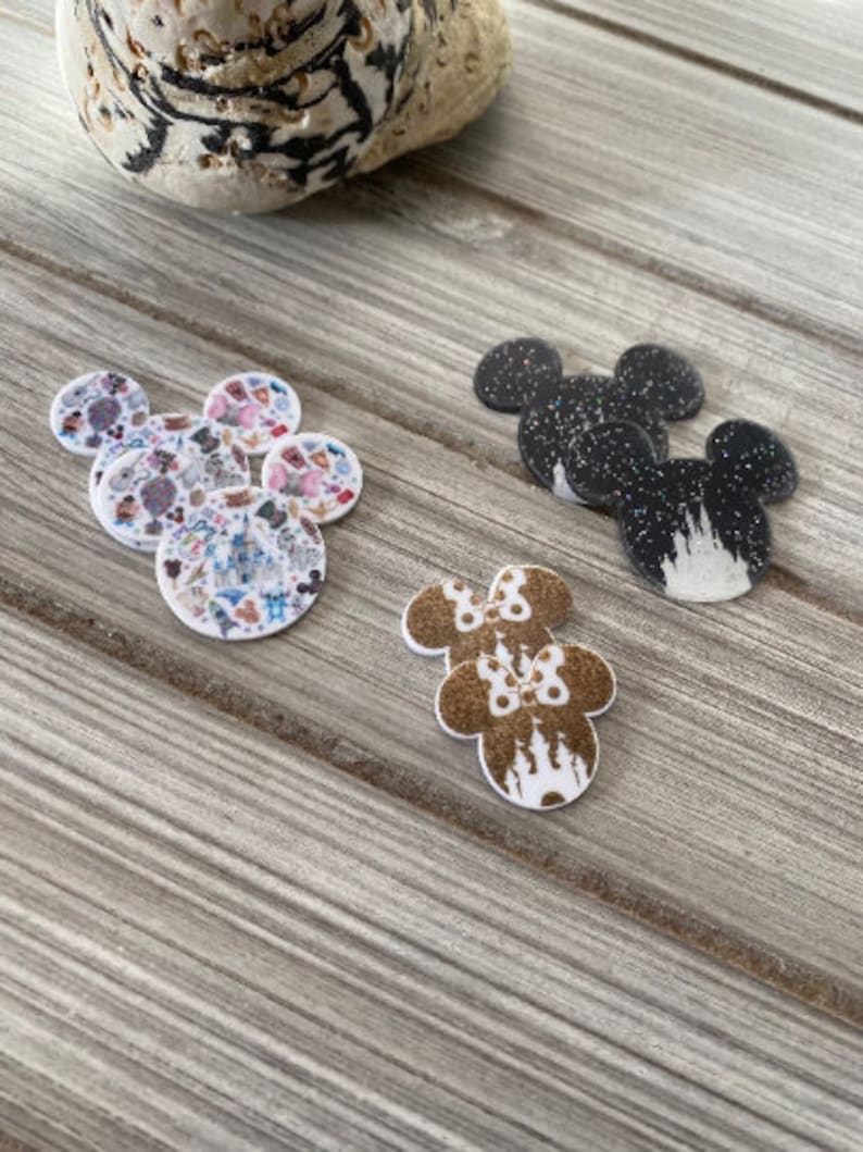 Puede incluir: Un conjunto de cinco adornos de resina en forma de orejas de rat&oacute;n con diferentes dise&ntilde;os. Un conjunto presenta un dise&ntilde;o de purpurina negro y blanco con una silueta de castillo blanco. Otro conjunto presenta un dise&ntilde;o de purpurina marr&oacute;n con una silueta de castillo blanco y un lazo rosa. El &uacute;ltimo conjunto presenta un dise&ntilde;o multicolor con varios personajes e iconos de Disney.