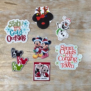 Puede incluir: Un conjunto de nueve pegatinas con temática navideña con Mickey Mouse y otros personajes de Disney. Las pegatinas incluyen Mickey Mouse, Minnie Mouse, un muñeco de nieve, Papá Noel y un reno. Las pegatinas son todas blancas con diseños y texto coloridos. El texto en las pegatinas incluye "Baby It's Cold Outside", "Ho Ho Ho", "Santa Claus is Coming to Town" y "Merry Christmas".