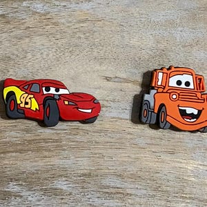 May include: Two red and orange cartoon car figurines. The red car is a race car with the number 95 on the side. The orange car is a tow truck with a smiling face.