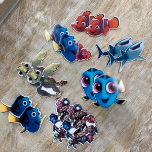 Puede incluir: Un conjunto de nueve parches termoadhesivos con temática de Buscando a Nemo. Los parches presentan personajes como Dory, Nemo, Marlin y un tiburón. Los parches también incluyen un ancla con el texto "Just Keep Swimming".