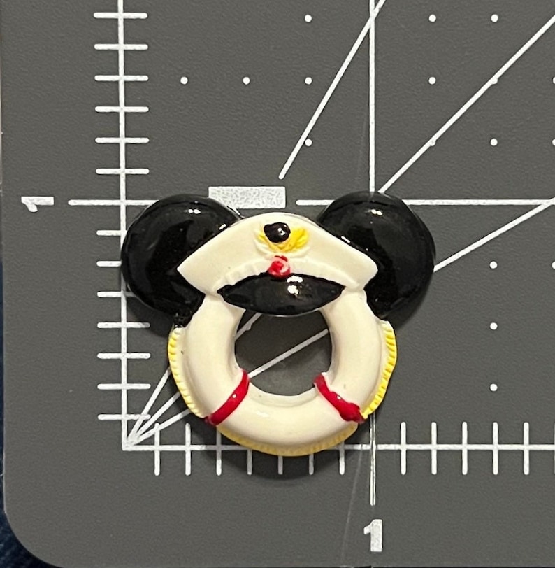 May include: A white ceramic life preserver with red trim and a black and white captain's hat with a yellow star on top. The life preserver has black mouse ears.