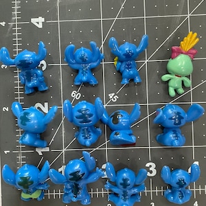 May include: A collection of 11 blue Stitch figurines, some with different poses and expressions. The figurines are made of plastic and have a glossy finish. One figurine is green and has a flower on its head.