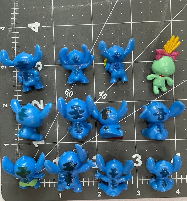 May include: A collection of 11 blue Stitch figurines, some with different poses and expressions. The figurines are made of plastic and have a glossy finish. One figurine is green and has a flower on its head.