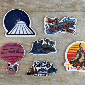 Puede incluir: Seis pegatinas de vinilo de colores que representan varias atracciones de los parques de Disney. Las pegatinas incluyen un Spaceship Earth, un globo aerostático con el texto "Walt Disney's It's a Small World", una montaña con el texto "Frontierland", una cascada con palmeras, una montaña con el texto "The Mountains Are Calling and I Must Go", y una ola con el texto "Have a Zip-A-Dee-Doo-Dah Day!"