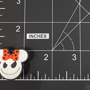 May include: A white and black shoe charm featuring a smiling character face with a black mouth and eyes, and a red bow with black polka dots. The charm is approximately 5 cm in length.