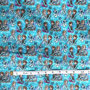 K&ouml;nnte beinhalten: Hellblauer Stoff mit einem sich wiederholenden Muster von Zeichentrickfiguren, darunter zwei junge Frauen und ein Schneemann. Die Figuren sind in verschiedenen Posen vor einem Hintergrund aus wei&szlig;en Schneeflocken. Der Stoff wird mit einem Ma&szlig;band gemessen.