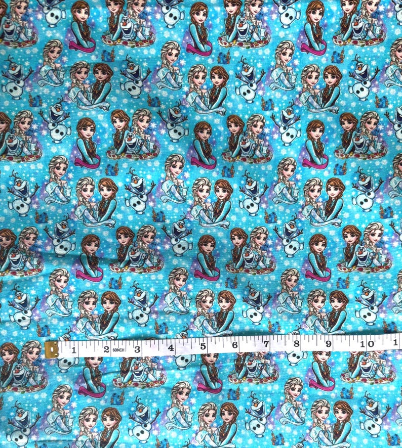 K&ouml;nnte beinhalten: Hellblauer Stoff mit einem sich wiederholenden Muster von Zeichentrickfiguren, darunter zwei junge Frauen und ein Schneemann. Die Figuren sind in verschiedenen Posen vor einem Hintergrund aus wei&szlig;en Schneeflocken. Der Stoff wird mit einem Ma&szlig;band gemessen.