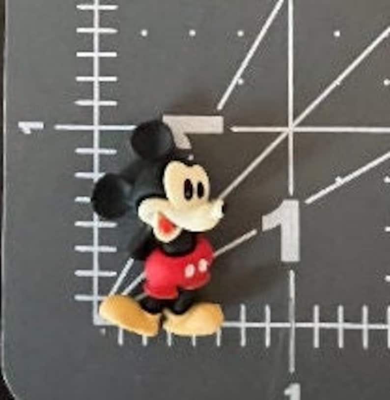 Puede incluir: Una peque&ntilde;a figura tridimensional de Mickey Mouse. La figura tiene cabeza y orejas negras, cara blanca, pantalones cortos rojos, zapatos amarillos y cuerpo negro. La figura mide aproximadamente 2,5 cm de altura.