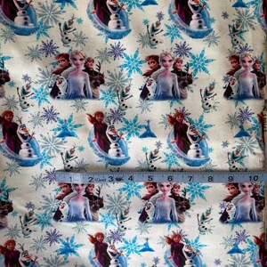 May include: White fabric featuring a repeating pattern of characters from the animated film "Frozen", including Elsa, Anna, Olaf, and snowflakes in shades of blue and purple. The fabric is measured with a ruler.