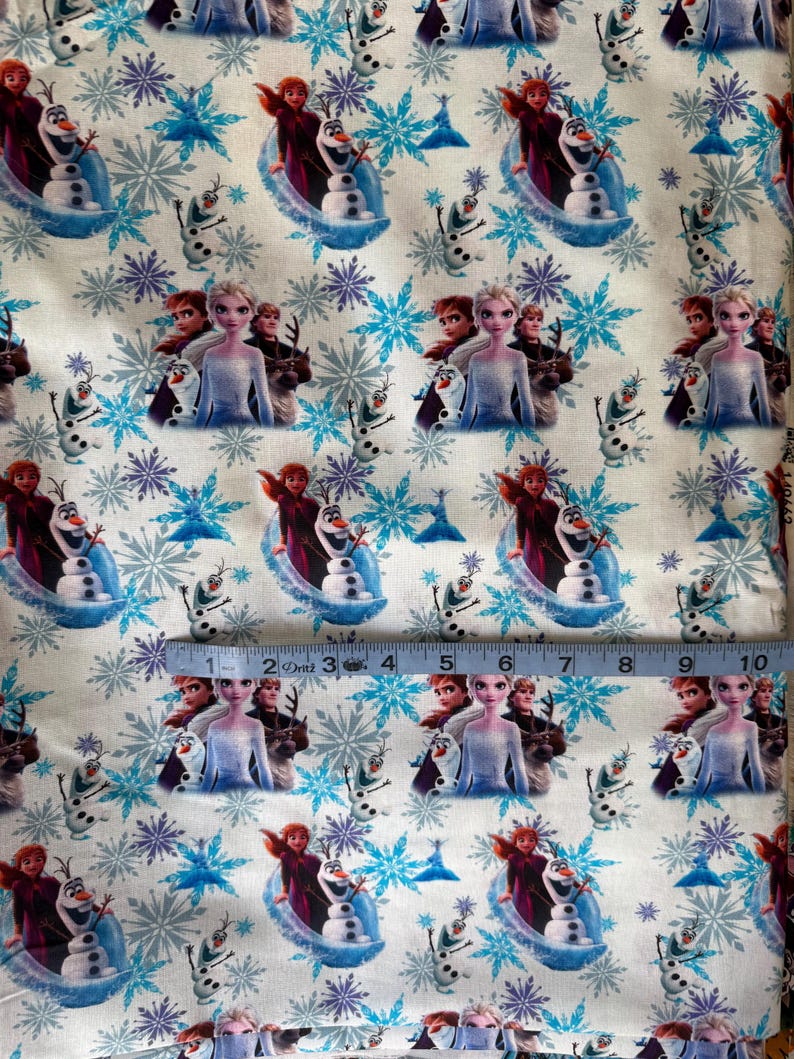 May include: White fabric featuring a repeating pattern of characters from the animated film "Frozen", including Elsa, Anna, Olaf, and snowflakes in shades of blue and purple. The fabric is measured with a ruler.