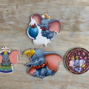 Könnte beinhalten: Vier bunte Aufkleber mit Dumbo dem Elefanten aus Disneys Dumbo. Die Aufkleber zeigen Dumbo in verschiedenen Posen, darunter fliegend mit einem Ballon, sitzend auf einer Zirkusplattform und in einem Glasmalereidesign.