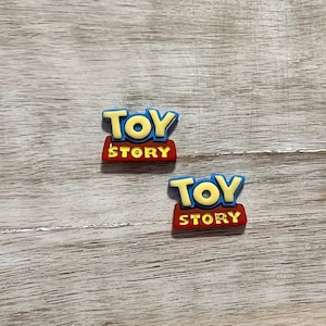 May include: Two blue and yellow Toy Story logo charms. The charms are made of rubber and have a red background.