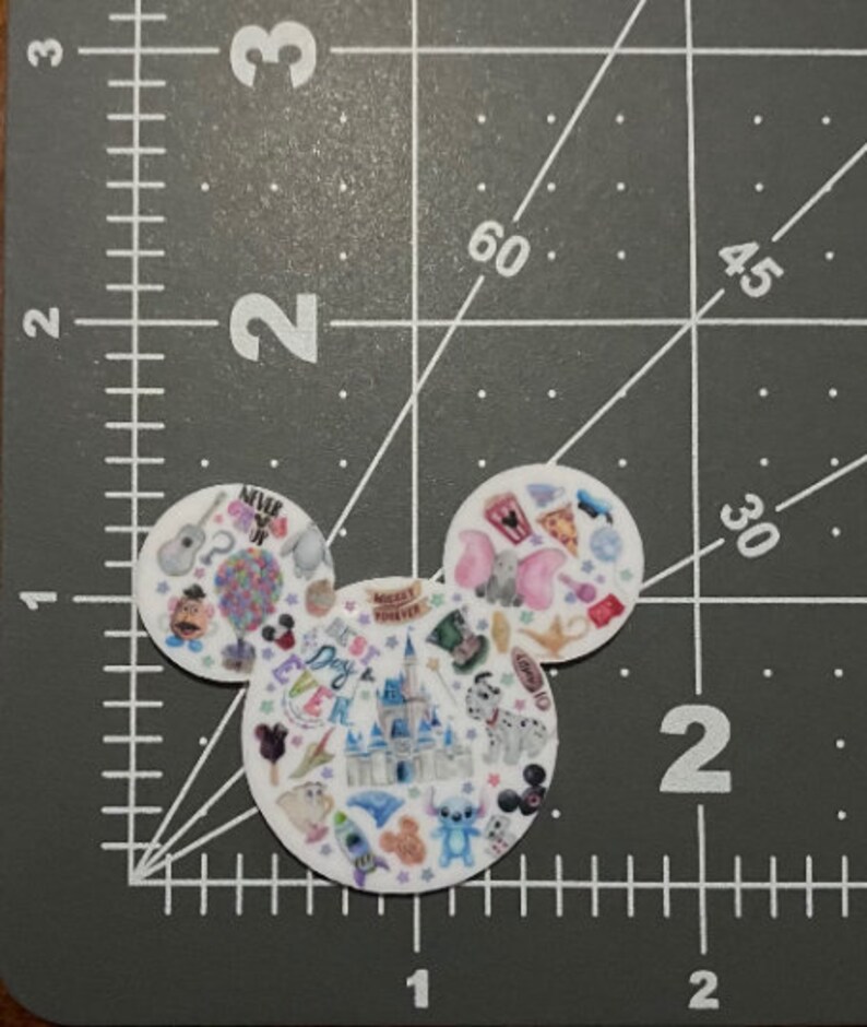 Puede incluir: Forma de cabeza de Mickey Mouse blanca con una variedad de personajes e iconos de Disney coloridos, incluidos Mickey Mouse, Minnie Mouse, Stitch y el castillo de Disney. El texto "Best Day Ever" es visible en el centro de la imagen.