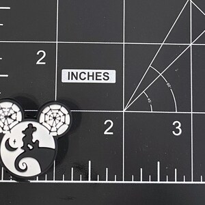 K&ouml;nnte beinhalten: Ein schwarz-wei&szlig;er Mickey Mouse-f&ouml;rmiger Charm mit Halloween-Motiv. Der Charm zeigt eine Silhouette einer Figur auf einem H&uuml;gel, einen Mond und Spinnweben in den Ohren. Der Charm ist etwa 5 cm gro&szlig;.