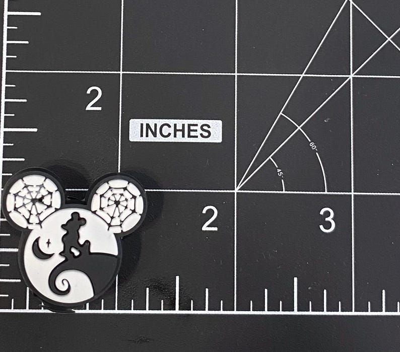 K&ouml;nnte beinhalten: Ein schwarz-wei&szlig;er Mickey Mouse-f&ouml;rmiger Charm mit Halloween-Motiv. Der Charm zeigt eine Silhouette einer Figur auf einem H&uuml;gel, einen Mond und Spinnweben in den Ohren. Der Charm ist etwa 5 cm gro&szlig;.