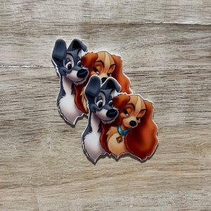 May include: Two die-cut stickers featuring the animated characters Tramp and Lady from the Disney movie, Lady and the Tramp. The stickers show the dogs in a cartoon style, with Tramp in gray and white, and Lady in tan and brown.