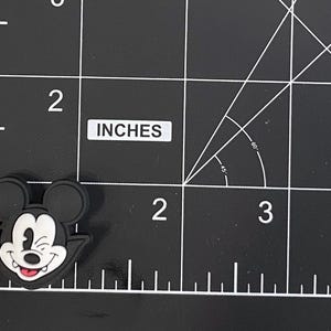 May include: A black and white Mickey Mouse-shaped charm with a winking expression and red tongue. The charm is approximately 3.8 cm in length. The charm is set against a black grid background with the word "INCHES" visible.
